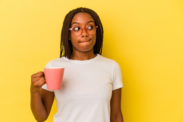 Young african american woman holding a mug isolated on yellow background  confused, feels doubtful and unsure.