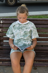 Teen girl sitting in the park and reading a book on a warm summer evening. Leisure of teenagers without a telephone.