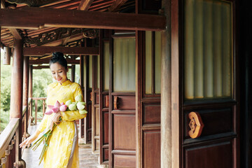 Fototapeta na wymiar Beautiful smiling young woman in ao dai dress standing on porch with bunch of lotus flowers