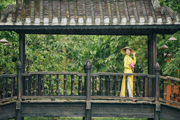 Smiling beautiful young Vietnamese woman in yellow dress and hat standing on bridge with bunch of lotus flowers and looking away