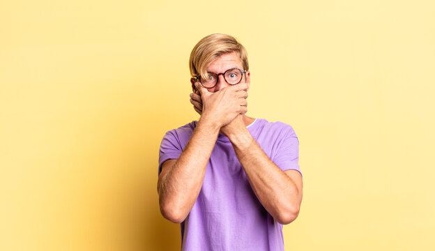 handsome blond adult man covering mouth with hands with a shocked, surprised expression, keeping a secret or saying oops
