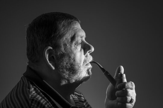 Black and white low key portrait of Caucasian bearded man smoking tobacco pipe against dark background