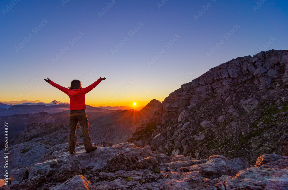 Wall mural happy hiker girl meets sunrise with raised arms high in the mountains.