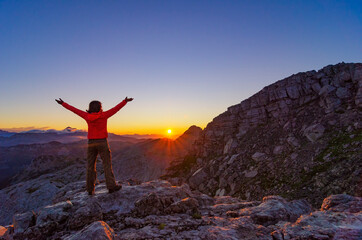 Happy hiker girl meets sunrise with raised arms high in the mountains.