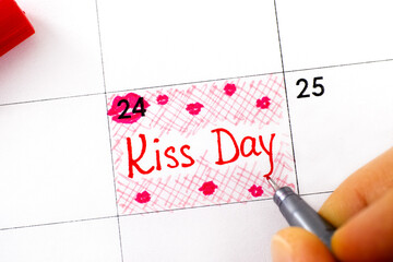 Woman fingers with pen writing reminder Kiss Day in calendar.