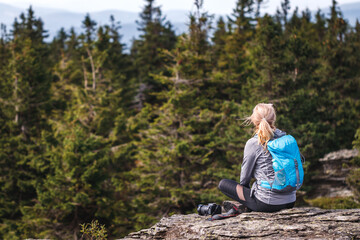 Woman with backpack and camera sitting on rock and enjoying view at forest. Tourist resting and relaxing at mountain peak