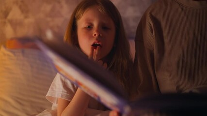Little girl puts fingers to mouth and mother takes away hand while reads tales book in bed at night lamp light in semidark children room closeup slow motion