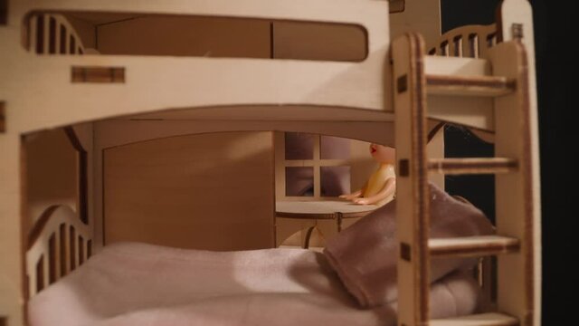Little girl suffering from cold peeks through window of miniature wooden house with doll at table and bunk bed in dark room closeup zoom in