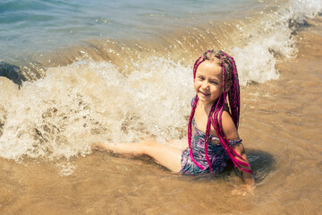 A girl with pink pigtails lies on the beach and smiles. Her feet are washed in waves.