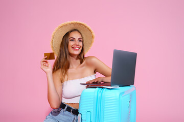  girl with laptop tickets, credit card and passport are going to travel. Sitting near suitcase in shorts white top and straw hat. Pink background. looking at laptop going to book something