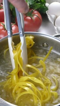 Chef cooking pasta in saucepan. Preparation italian tagliatelle in domestic kitchen. Cook with tongs mixing pasta in saucepan. Mushrooms, eggs, tomatoes. Vertical video.