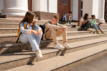 Two schoolkids in casualwear discussing plans for the day while sitting on large staircase