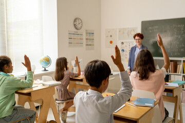 Rear view of group of intercultural classmates sitting by desks and raisng hands at lesson