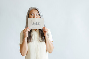 Shocked silver haired Asian lady holds card with word Sale on light grey background in studio, space for text