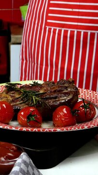 Chef decorates fried meat in plate with fried cherry tomatoes and rosemary. Vertical video. Close-up. Indoors.