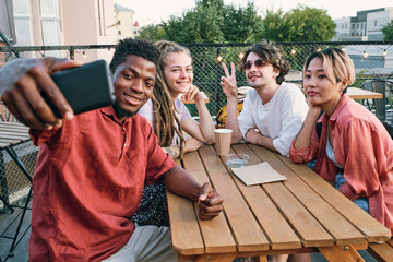 Happy African guy making selfie with intercultura friends by wooden table in urban environment