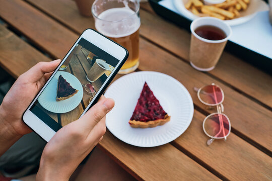 Hands of young female with smartphone taking photo of tasty cake on plate and drinks