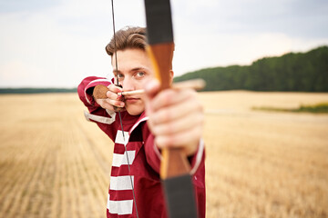 Young male sportsman targeting with bow in a traditional, medieval archer costume - Teenager archer practicing archery in nature - Field archery and recreation concept with a millennial boy