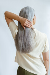 Middle aged woman in yellow blouse adjusts long loose hoary hair on light grey background in studio backside view. Mature beauty lifestyle