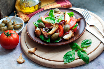 Italian Panzanella salad with bread, tomatoes, basil, capers and olive oil on round wooden board on gray background