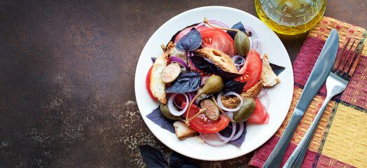Italian Panzanella salad with bread, red onion, tomatoes, basil, capers and olive oil on white plate on dark brown background. Top view