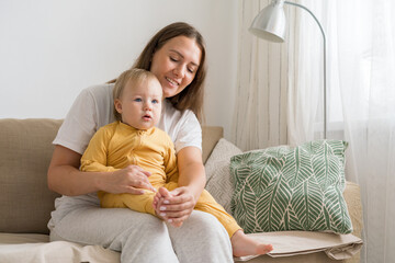 Smiling and happy young female in casual clothing sitting on couch with pretty blond blue-eyed baby...