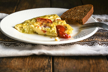 A slice of rustic omelet with potatoes and tomatoes on a white plate. A slice of bread, a kitchen napkin, and a wooden table.