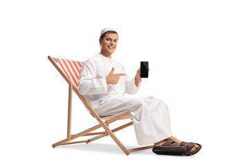Young man in ethnic clothes seated on a beach chair and pointing at a mobile phone