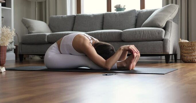 Young 30s athletic beautiful woman in white comfort sportive wear seated on mat in forward bend or Paschimottanasana intense dorsal stretch asana. Yoga practise at home, quarantine, lifestyle concept