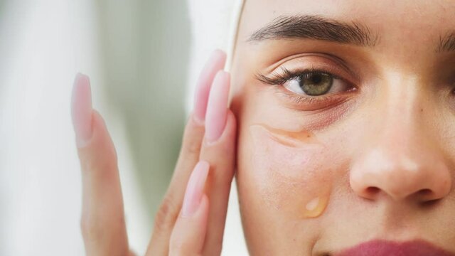 Woman applying face serum using pipette for moisturizing, morning skincare routine. Young female student wearing bathrobe looking in mirror in spa salon. Natural day make up. Half-face portrait.