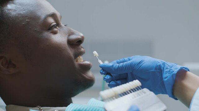 Dentist applying tooth scale samples set to smiling black man patient at dental clinic, close up portrait, slow motion