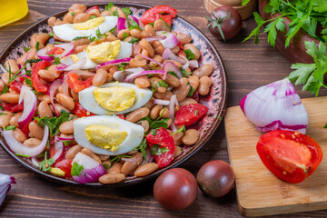 Wooden table with a plate with Turkish classiyechka snack for piyaz meat. Tomatoes, boiled eggs, white beans and onions.