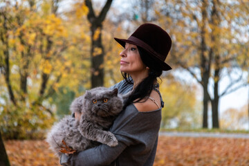 A girl in a hat holds a fluffy British cat against the backdrop of a yellow autumn forest.