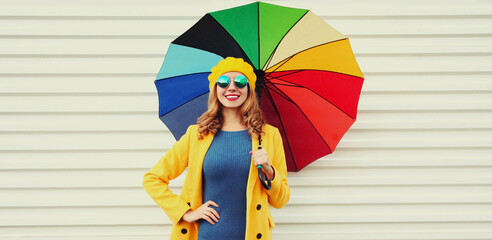 Autumn portrait of happy cheerful smiling young woman model with colorful umbrella wearing a yellow coat and beret on a white background