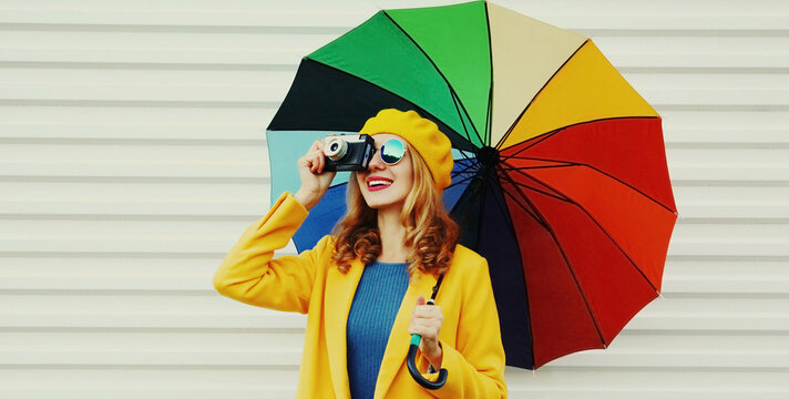 Autumn portrait of happy cheerful smiling young woman photographer with colorful umbrella and camera wearing a yellow coat and beret on a white background