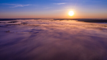 Fototapeta premium Aerial view of a beautiful summer landscape with a fog while dawn. Photo from drone of a foggy landscape in spring. Top view to land while sunrise. Sun is above the horizon