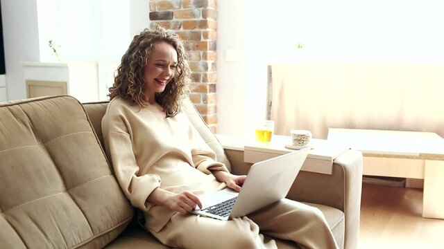Beautiful cheerful girl using silver laptop while sitting on sofa in living room at home. Attractive businesswoman studying online, using laptop software.