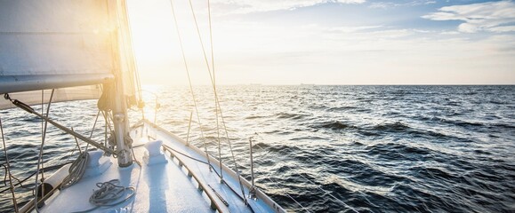 Yacht sailing in an open sea at sunset. Close-up view of the deck, mast and sails. Clear sky after the rain, glowing clouds, golden sunlight. Panoramic seascape - obrazy, fototapety, plakaty