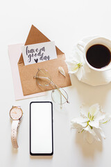 Inspirational quote: "have a good day" inside a kraft envelope. Home office morning composition with: mobile phone, wrist watch, white flowers, glasses and a cup of coffee. Flat lay, top view.