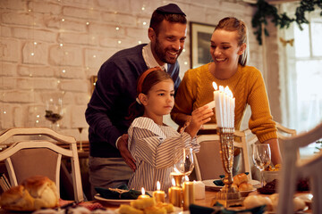 Happy Jewish family lights menorah candles before meal at dining table during Hanukkah.