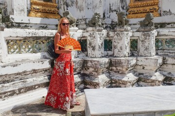 Travel to Asia. Portrait of young caucasian blonde girl traveler with traditional asian fan in hands who visiting famous buddhist Temple Wat Pho or Reclining Buddha Temple in Bangkok, Thailand