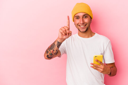 Young caucasian man holding mobile phone isolated on pink background  showing number one with finger.