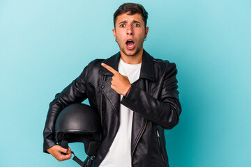 Young biker man holding helmet isolated on blue background  pointing to the side