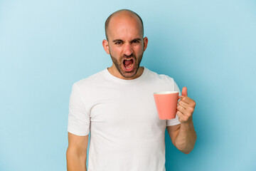 Young bald man holding a mug isolated on blue background  screaming very angry and aggressive.