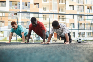 Positive black father teaching teenage sons to do push-ups on sports ground against flat block