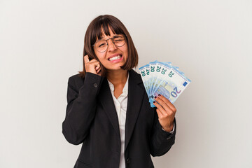 Young mixed race business woman holding a banknotes isolated on white background covering ears with hands.