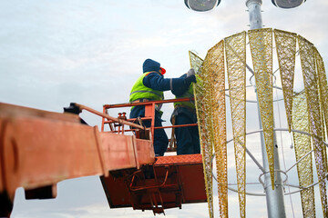 Workers install garlands and Christmas decoration on lamp post. Industrial climber decorate street light. Utility worker decorate street lamp for New Year. Men in lift bucket decorating street lamp