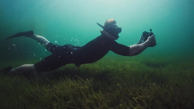Underwater photographer. Man in wetsuit slowly swims underwater with underwater camera in housing