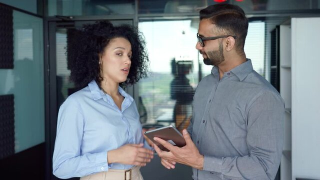 Smiling happy Indian male ceo businessman mentor and female African American manager intern discussing project using tablet device standing in modern corporate office. Business concept.