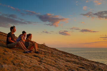 friends two women and a man at sunset time sit on a mountain above the sea and look into the distance